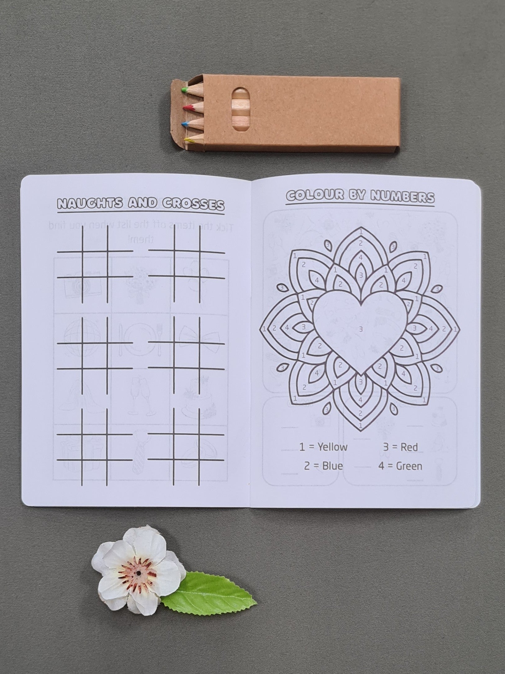 Inside pages of a personalised wedding activity booklet for kids, showing the 'Noughts and Crosses' and 'Colour by Number' activities. Coloured pencils included.